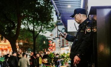 A street guard stands focused on his phone amid the bustling night streets of Nanjing, China.