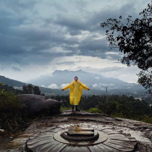 A person in a yellow raincoat standing on a stone sculpture in Wayanad, India, with a dramatic cloudy sky.