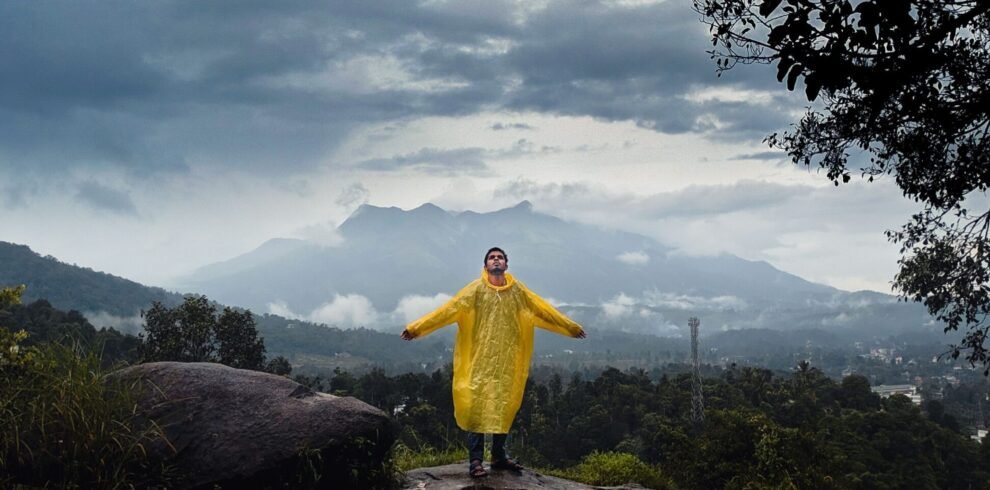 A person in a yellow raincoat standing on a stone sculpture in Wayanad, India, with a dramatic cloudy sky.