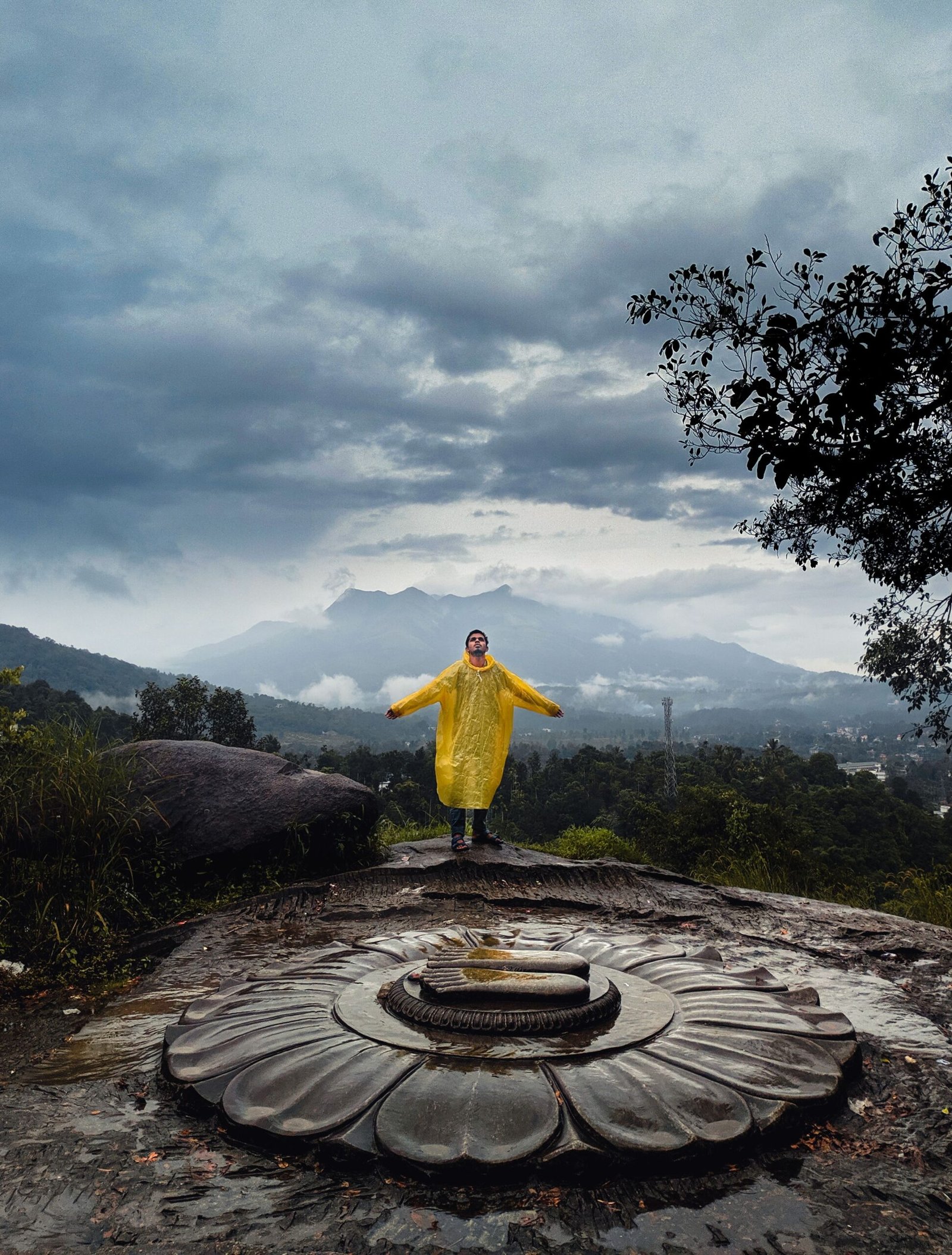 A person in a yellow raincoat standing on a stone sculpture in Wayanad, India, with a dramatic cloudy sky.