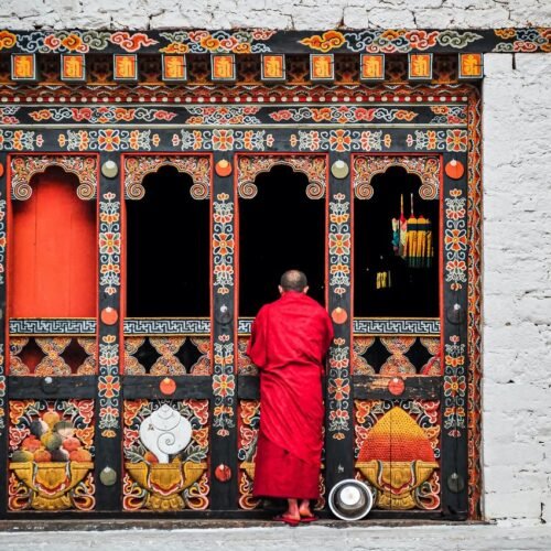 A Buddhist monk in red robes standing at an ornately decorated temple doorway in Bhutan.