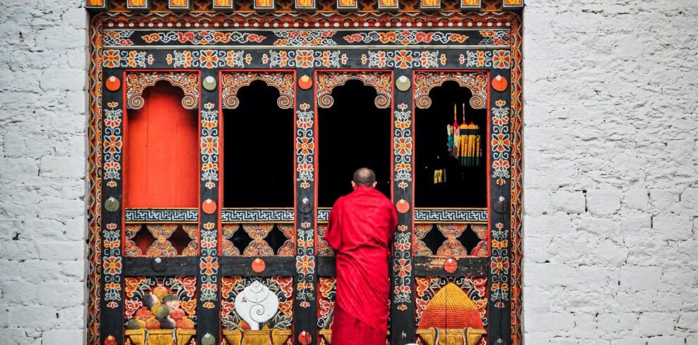 A Buddhist monk in red robes standing at an ornately decorated temple doorway in Bhutan.