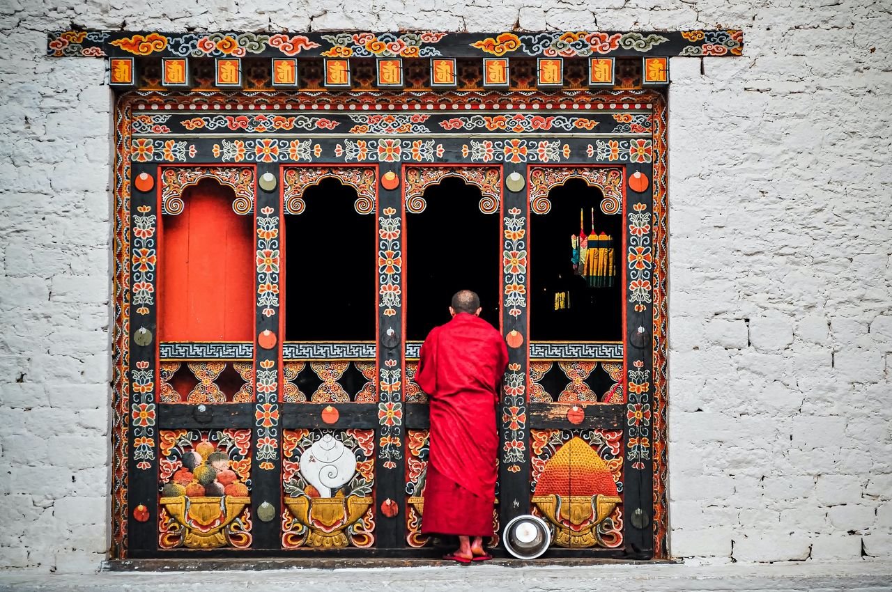 A Buddhist monk in red robes standing at an ornately decorated temple doorway in Bhutan.