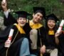 Three diverse graduates joyfully celebrating outdoors in caps, gowns, and holding diplomas.