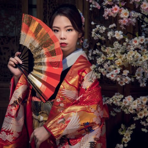 An Asian woman in a vibrant kimono poses gracefully with a fan against a backdrop of sakura blossoms.