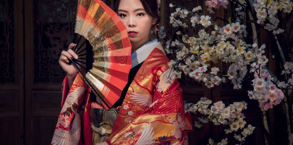 An Asian woman in a vibrant kimono poses gracefully with a fan against a backdrop of sakura blossoms.