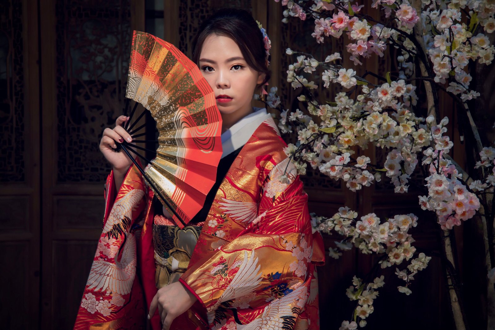 An Asian woman in a vibrant kimono poses gracefully with a fan against a backdrop of sakura blossoms.